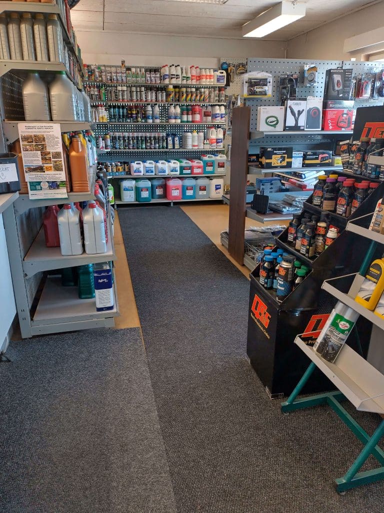 Kuva Laihian autotarvikeliikkeen sisätilasta, jossa on hyllyjä täynnä autojen huolto- ja kunnossapitotuotteita. Vasemmalla näkyy öljykanistereita eri kokoisissa ja värisissä pakkauksissa. Keskellä takaseinällä on laaja valikoima aerosoleja, pesuaineita, jäähdytinnesteitä ja muita kemikaaleja suurissa kanistereissa. Oikealla on hyllyssä DFC-merkkisiä lisäaineita ja autonhoitotuotteita. Lisäksi näkyy joitakin sähkötyökalupakkauksia ja kaapeleita. Lattialla on tummanharmaa matto, ja tilaa valaisee katossa oleva loisteputkivalaisin.